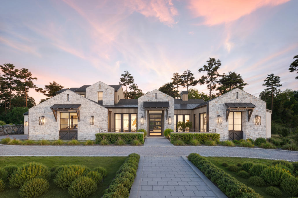 Modern house with stone exterior and large windows, surrounded by landscaped gardens, at sunset.