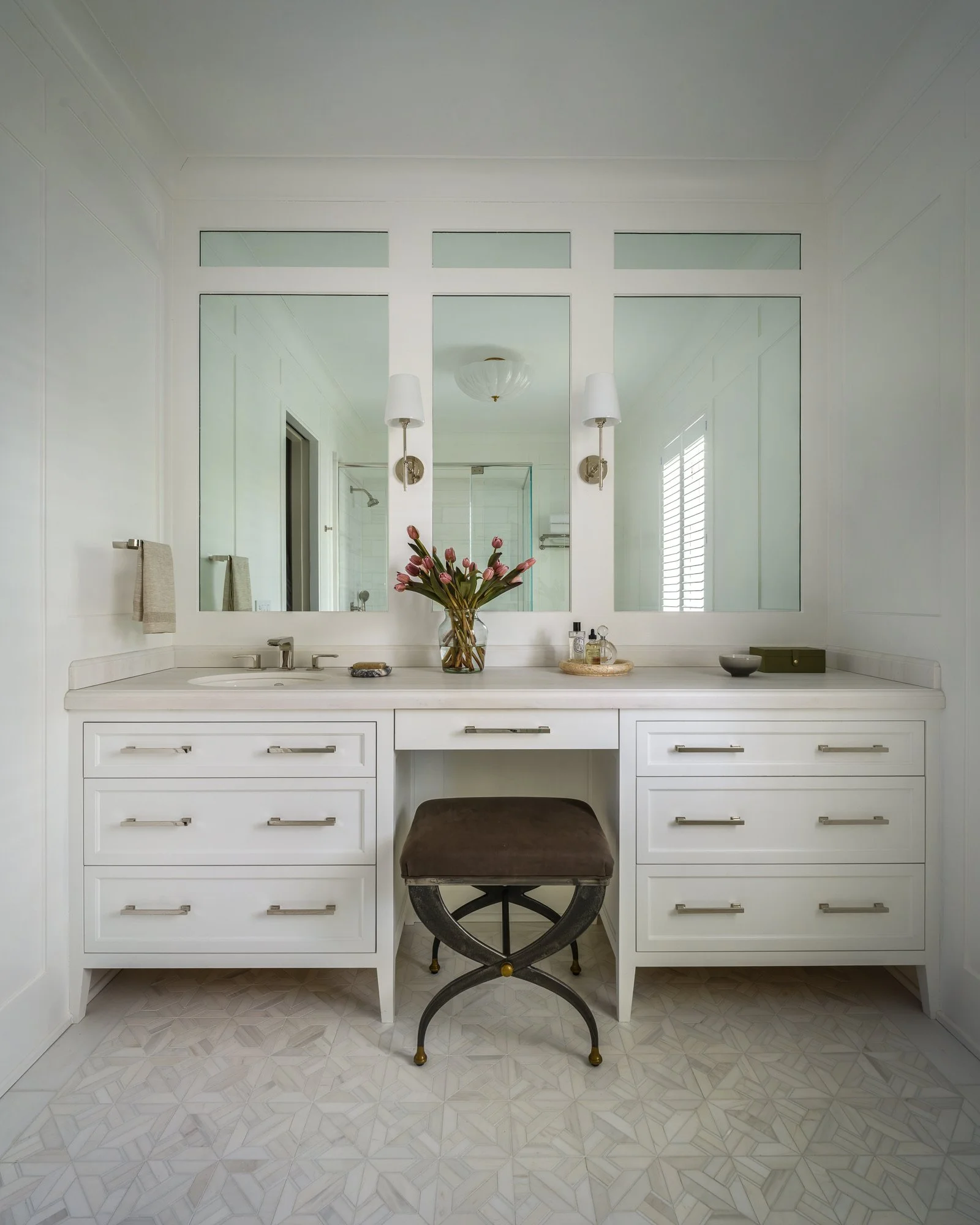Bathroom vanity with large mirror, white cabinets, and a stool with a dark cushion. Decor includes a vase of pink flowers, small bottles, and bowls. Lighting fixtures are mounted on the wall near the mirror.