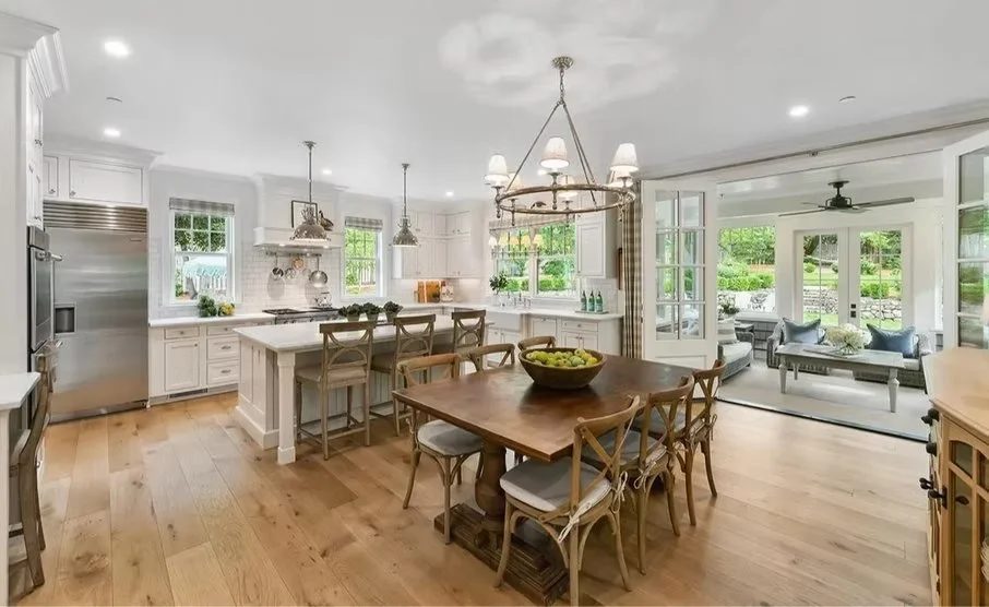 Open-concept kitchen and dining area with white cabinets, stainless steel appliances, wooden dining table with chairs, and a sunroom with seating visible through large glass doors.