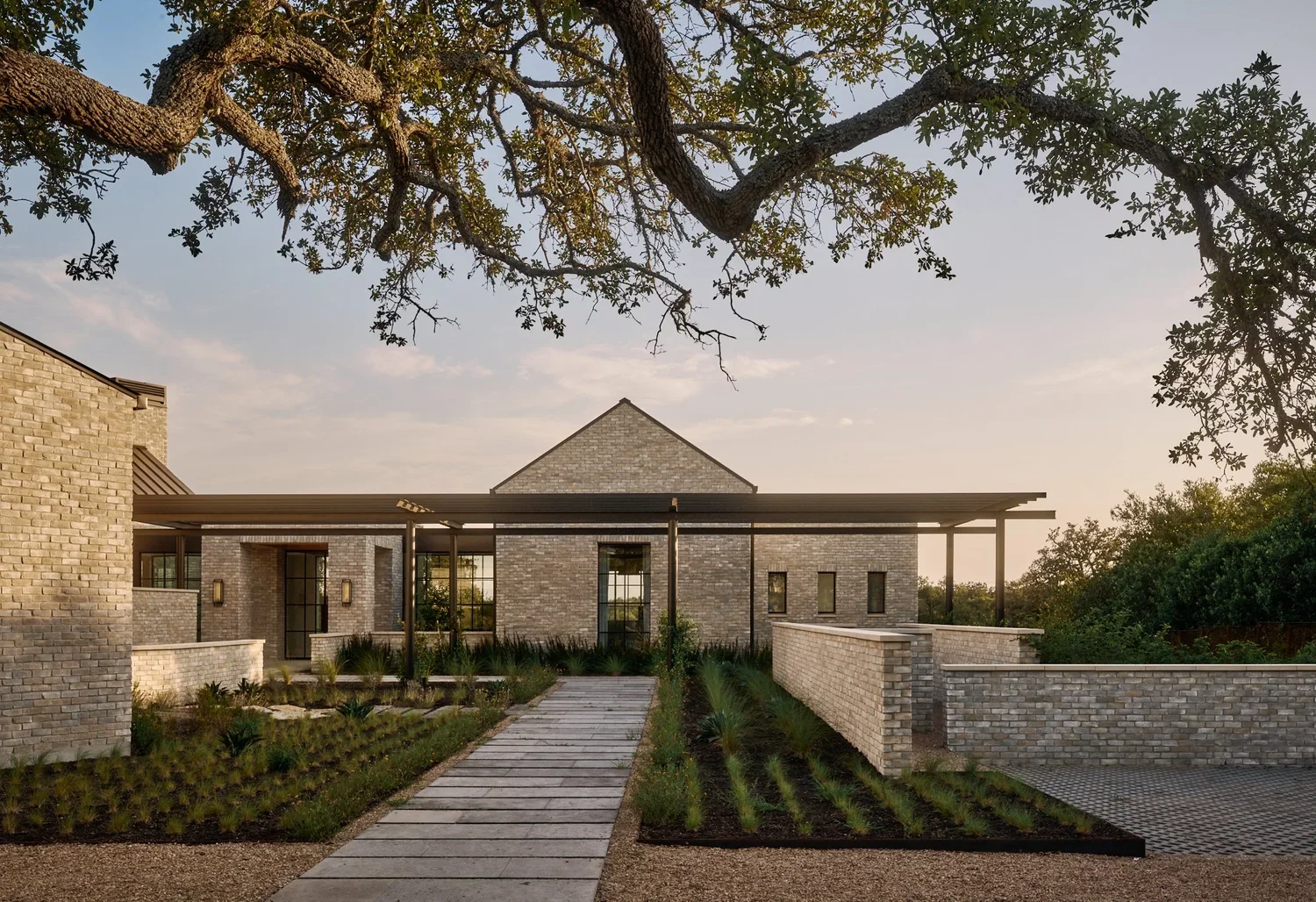 Modern brick house with a garden, pathway, and overhanging tree branches, during daylight.