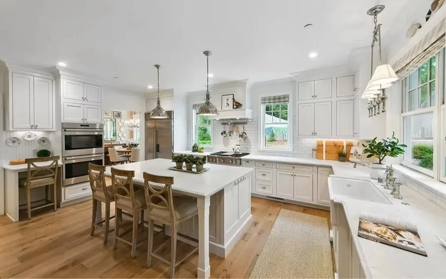 Bright, white kitchen with island, white cabinets, stainless steel appliances, multiple windows, wooden bar stools, and light hardwood flooring.
