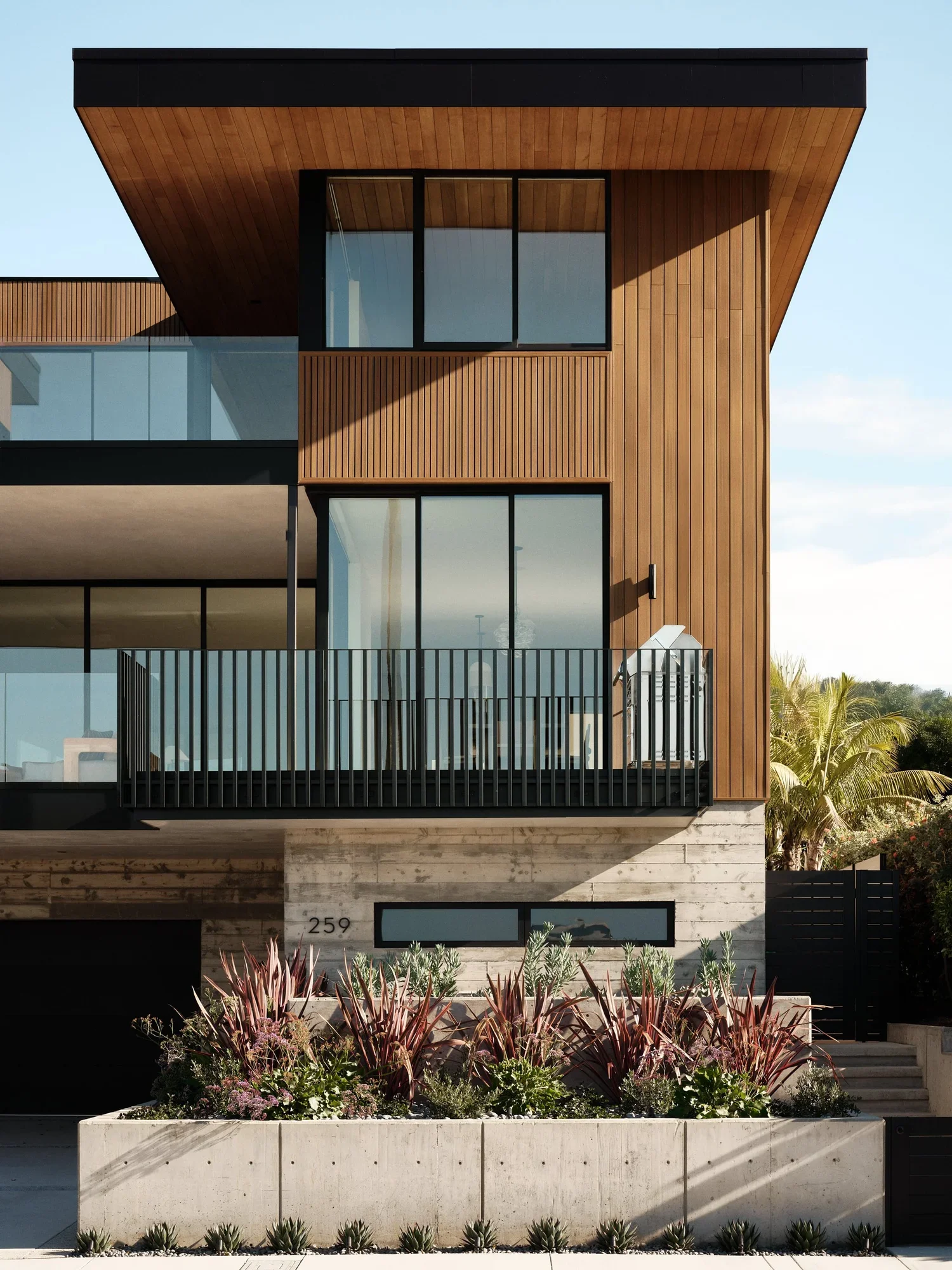 Modern building with wood paneling, large glass windows, and a balcony, with landscaped plants in front.