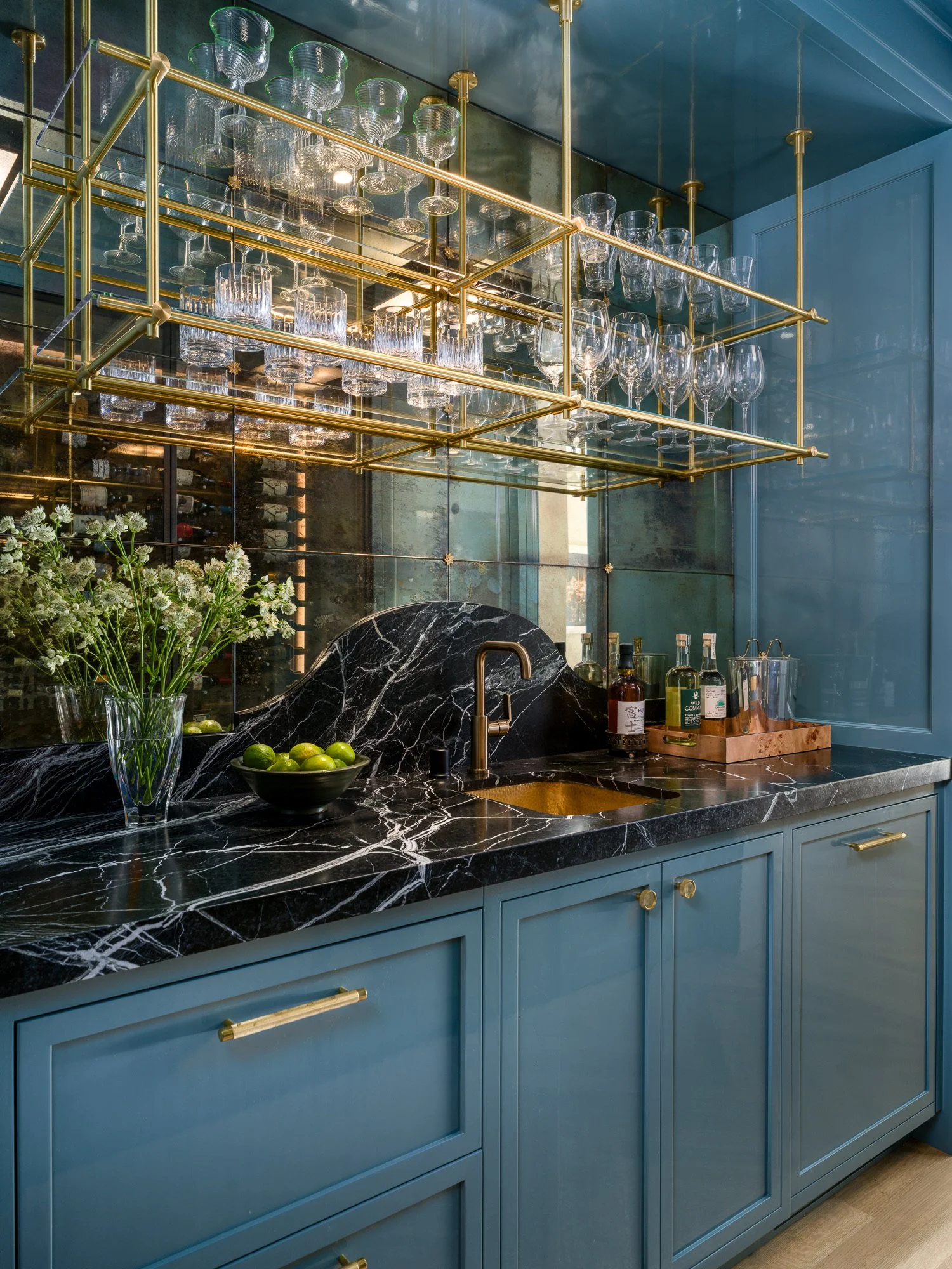 Kitchen with blue cabinets, black marble countertop, gold hardware, glassware on gold racks, and a black marble backsplash.