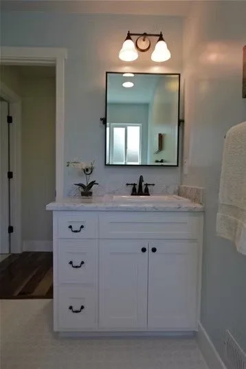 White bathroom vanity with a marble countertop, three drawers, two cabinets, and a black faucet, reflected in a wall-mounted mirror with dual light fixtures above.