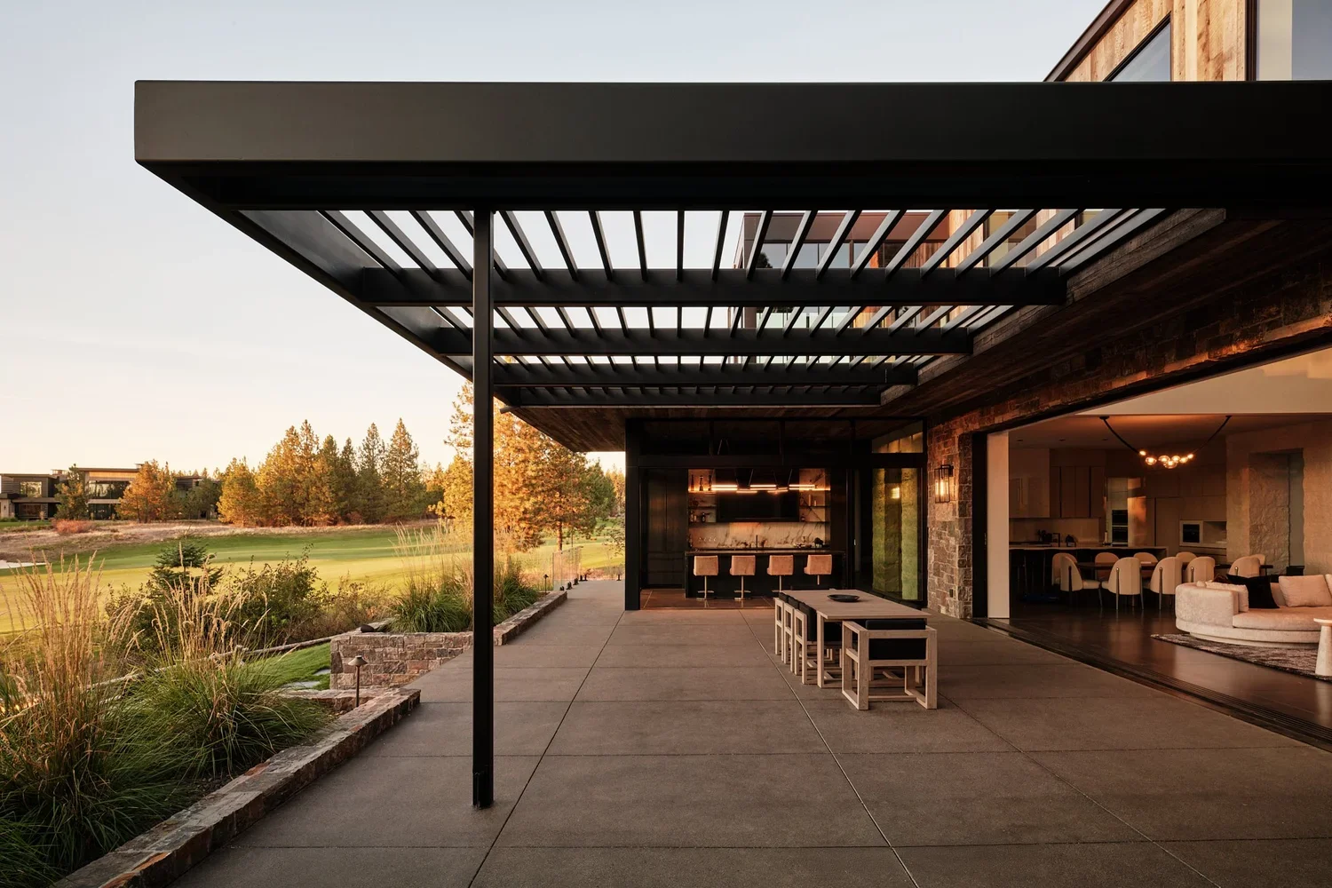 Modern outdoor patio area with black pergola, concrete flooring, and views of a green landscape with trees.