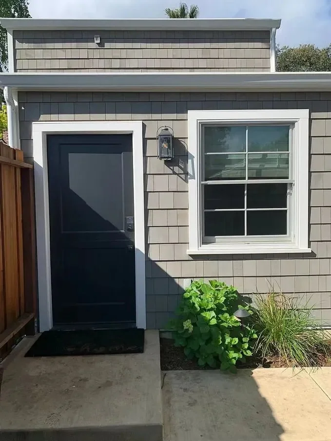 Exterior of a house with gray shingle siding, a black door with a white frame, a window with white framing, and a small garden with green plants near the door. There is a black outdoor light fixture beside the door.