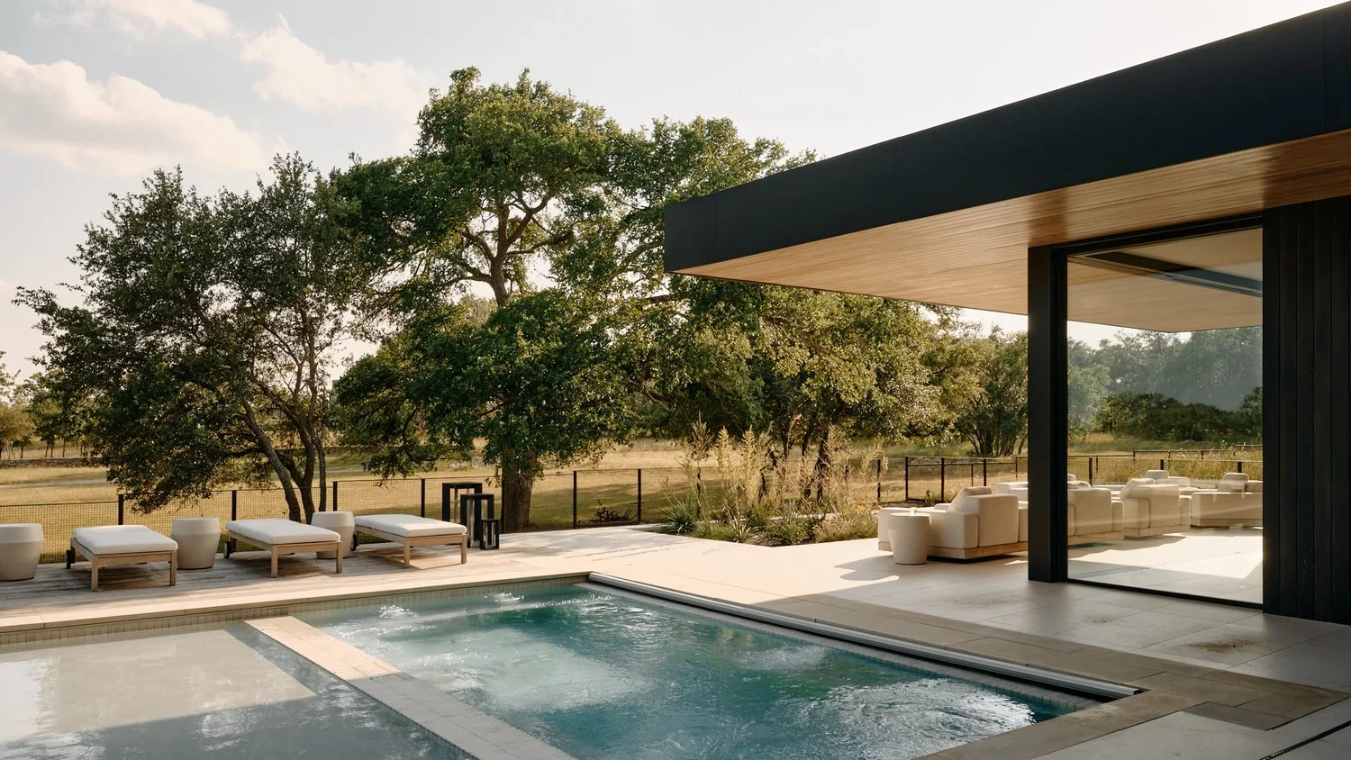 Modern backyard with a rectangular pool, sun loungers, white cushioned outdoor furniture, trees, and a patio under a house extension in a rural setting.