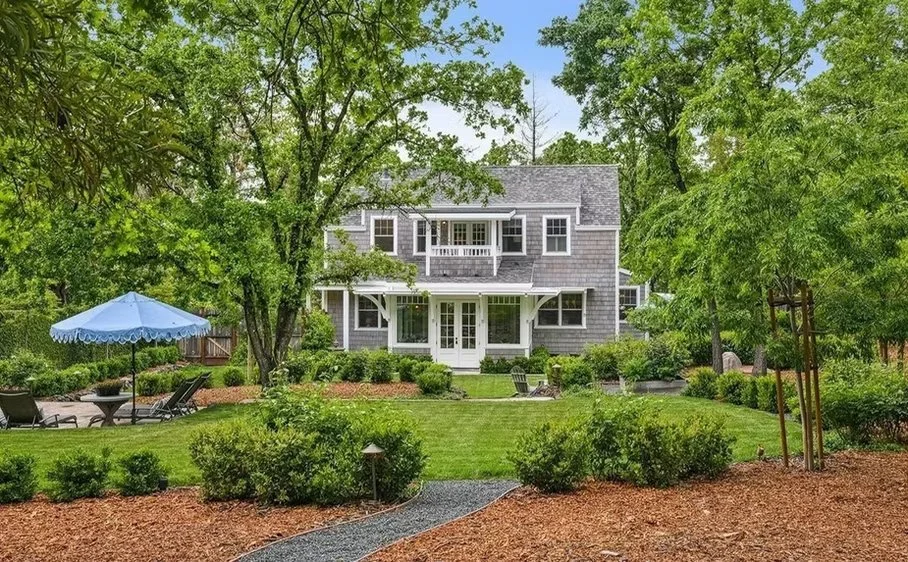 A spacious backyard with green grass, shrubs, and trees. There are lounge chairs and a blue umbrella on the left side, and a gray house with two stories and large windows in the background.
