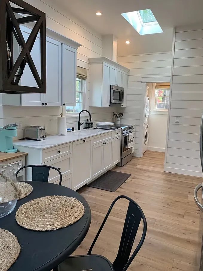 A modern kitchen with white cabinets, a black faucet, stainless steel appliances including a microwave and oven, a round black dining table with woven placemats, black chairs, a skylight, wooden flooring, and a small laundry area in the background.