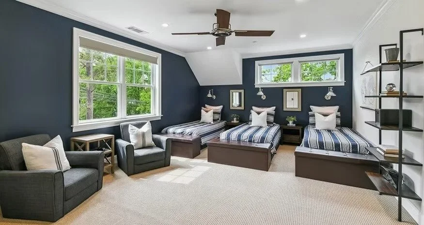 A bedroom with three twin beds with striped bedding, a dark blue accent wall, two large windows, a ceiling fan, and modern black shelving on the wall.