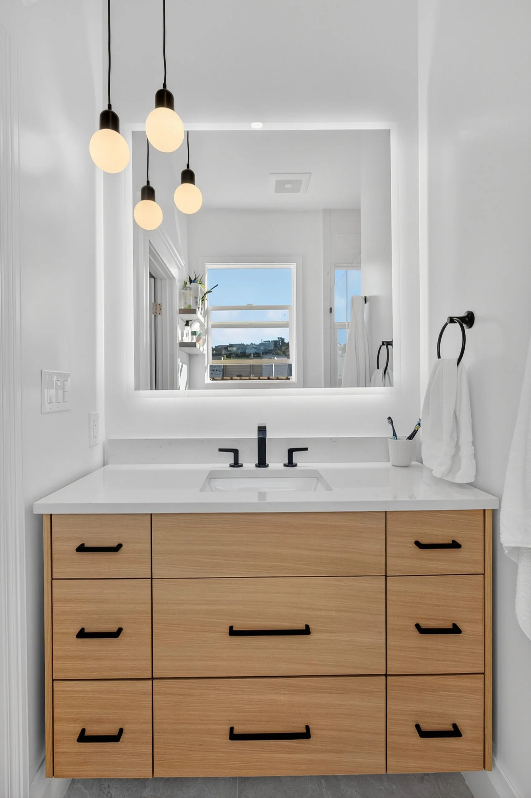 A bathroom vanity with a white countertop, a black faucet, and a wooden cabinet with black handles. Behind the vanity, there is a large mirror reflecting a window with a view of buildings and a blue sky. To the right, white towels are hanging on blac