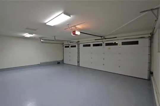 Empty two-car garage with white painted walls, gray floor, and a large sectional garage door. Ceiling has fluorescent lighting and garage door openers.