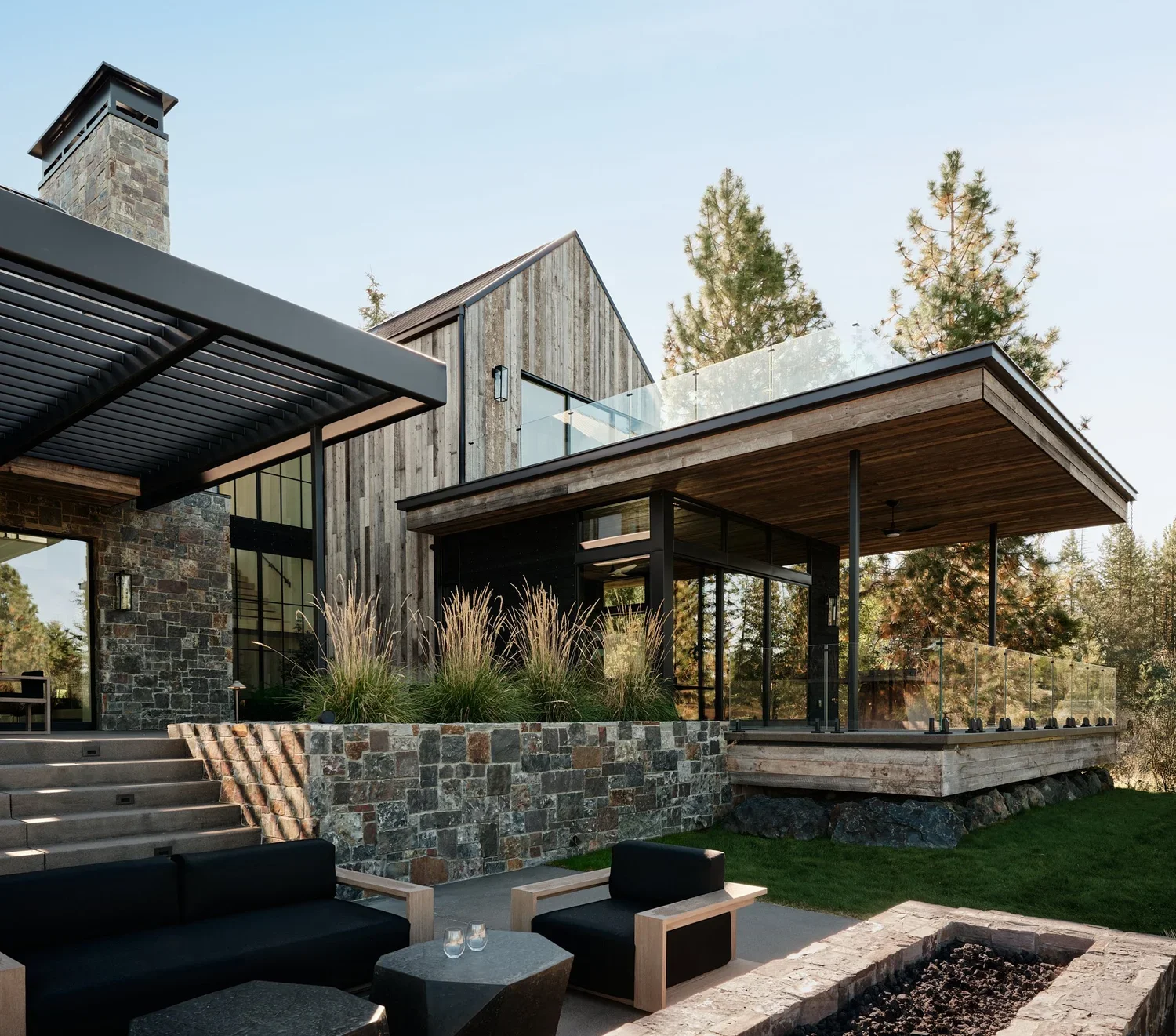 Modern house with stone and wooden exterior, large glass windows, and an outdoor seating area with black and wooden furniture, surrounded by tall trees.