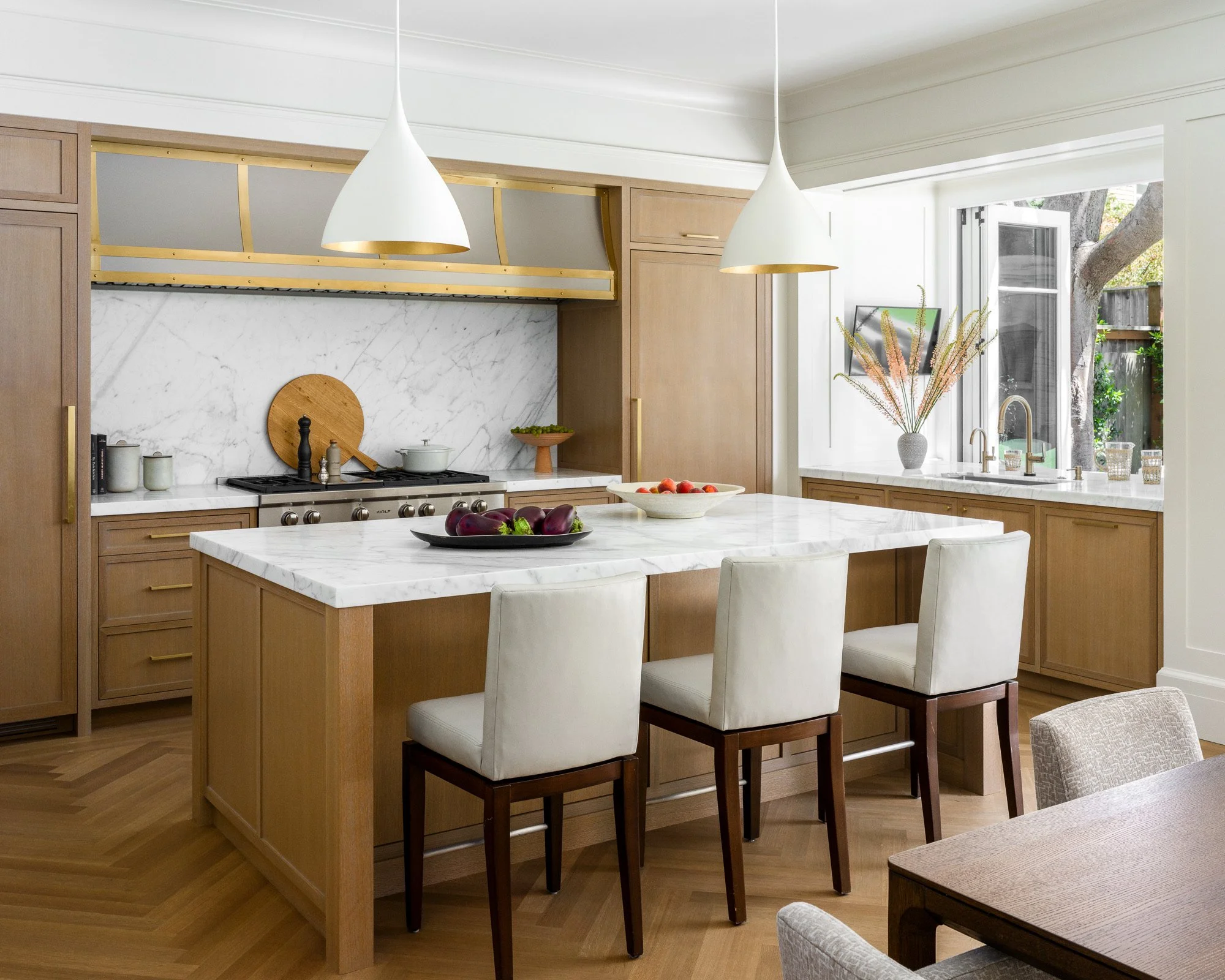Modern kitchen with light wood cabinets, white marble countertops, and stainless steel appliances. There is a kitchen island with white chairs, a bowl of fresh vegetables, and a window with a view of a tree outside.
