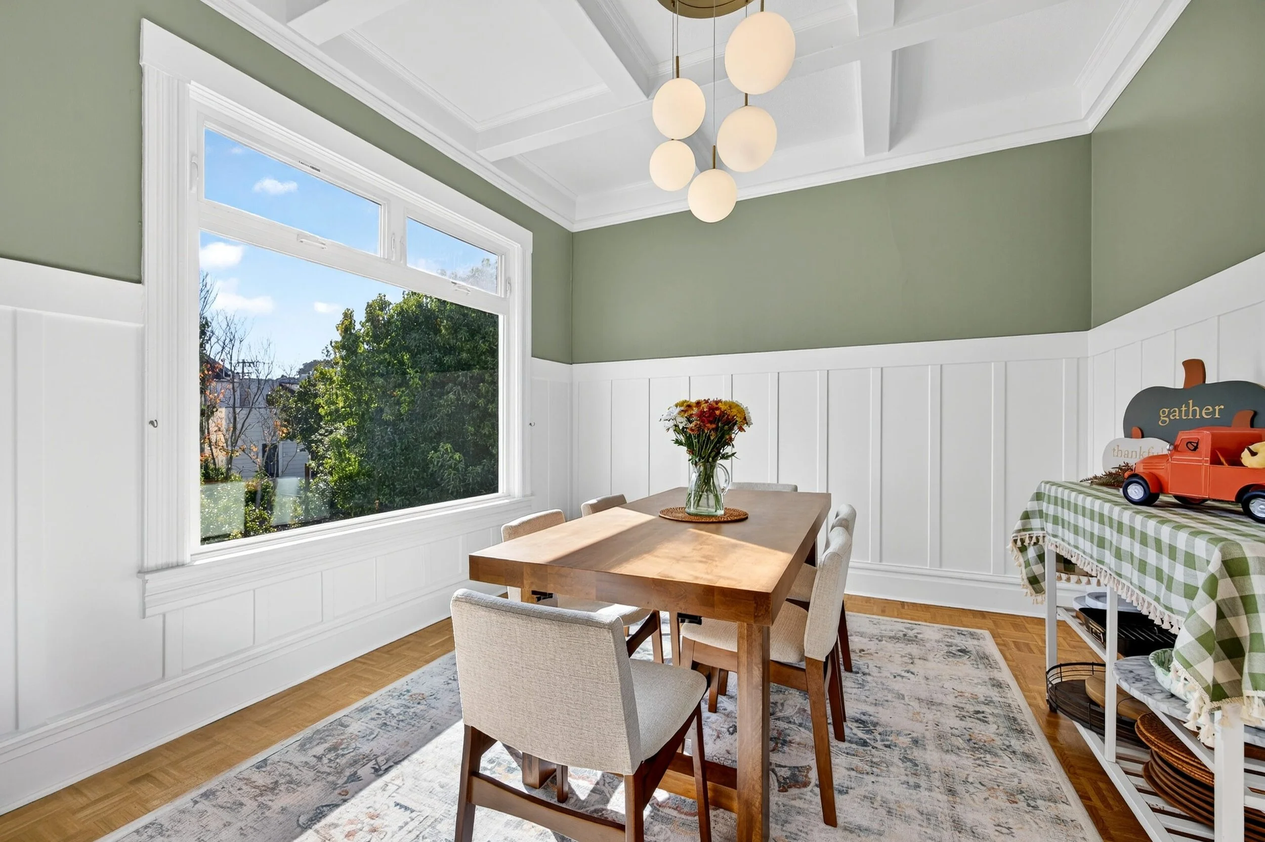 A dining room with a wooden table, four beige chairs, a vase of flowers, white wainscoting, a large window showing trees outside, a patterned rug, a shelf with a red toy fire truck, and a modern chandelier with six round white lights.