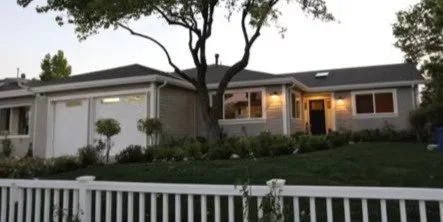 Front view of a single-story house with a white fence, a large tree in the yard, and exterior lights on at dusk.