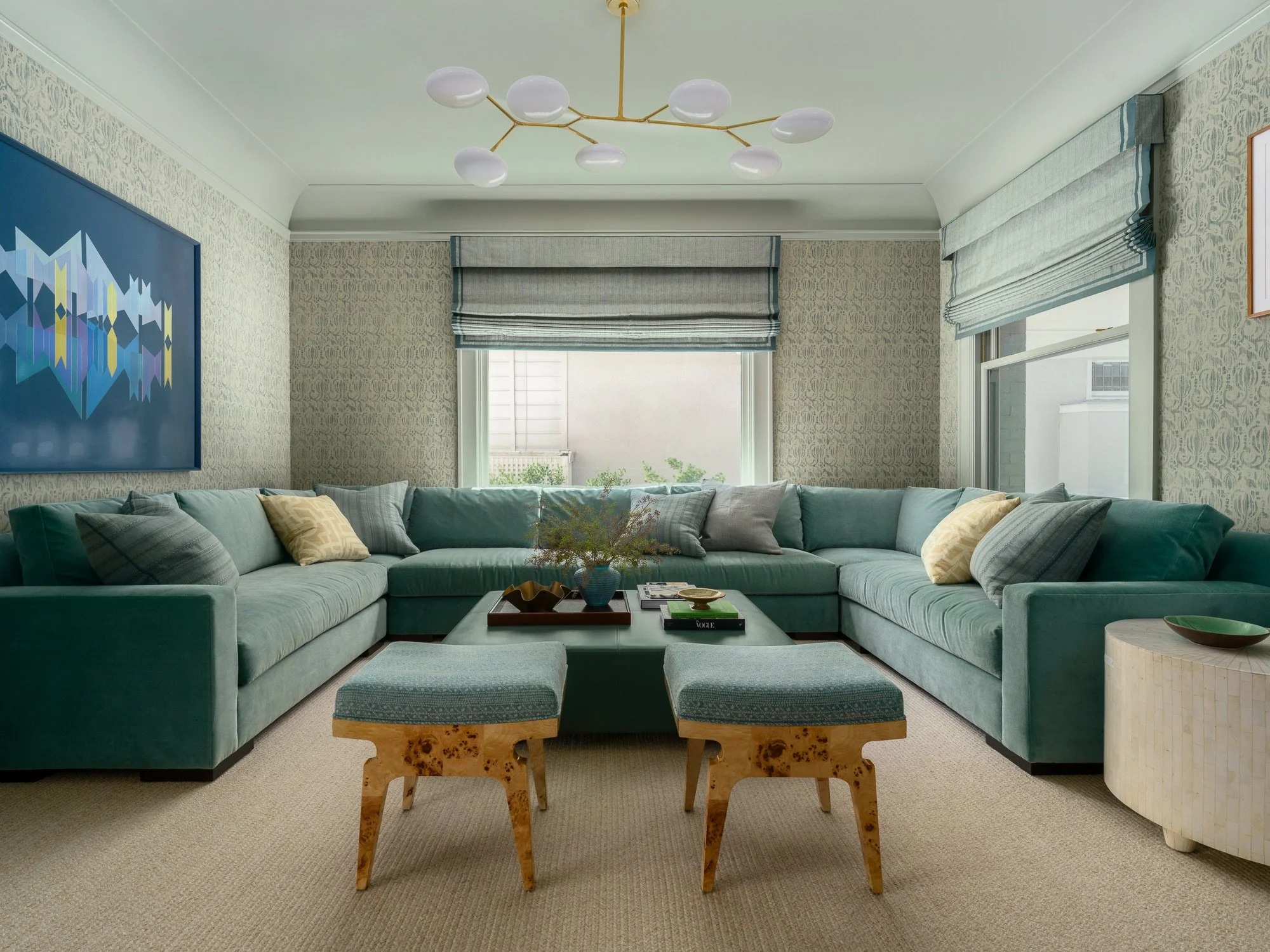 A cozy living room with teal sectional sofa, patterned throw pillows, wooden coffee table with decor, wall art, patterned wallpaper, large windows with striped roman shades, and ceiling light fixture with multiple white globes.