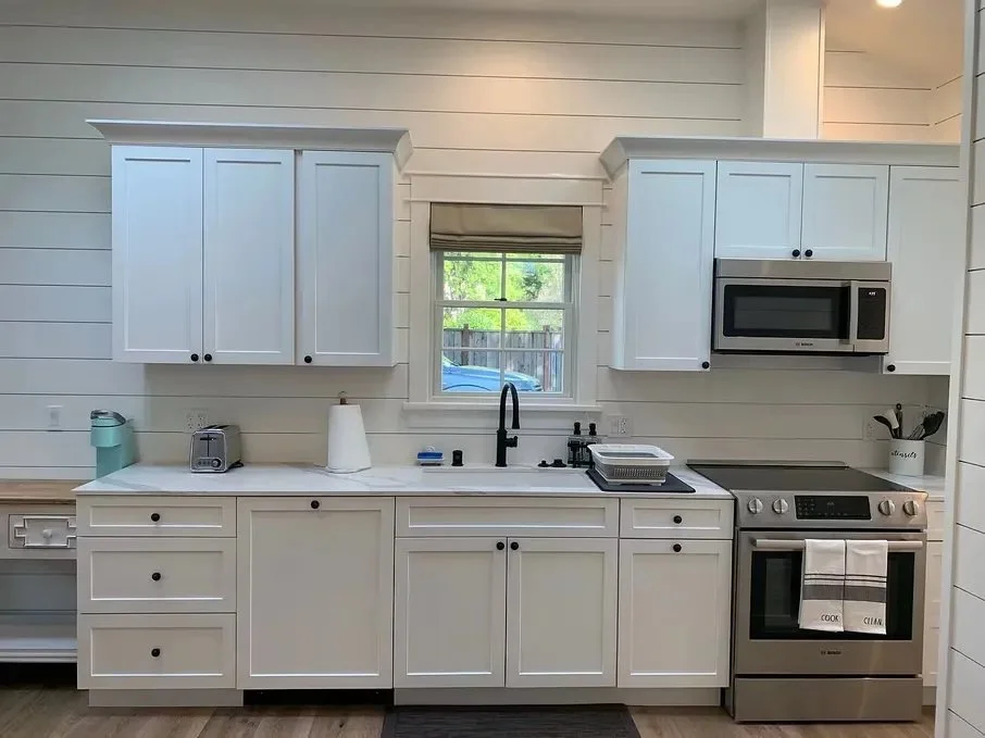 White kitchen with upper and lower cabinets, a small window above the sink, and stainless steel appliances including a microwave and oven. Countertop has a paper towel roll, toaster, and utensils.