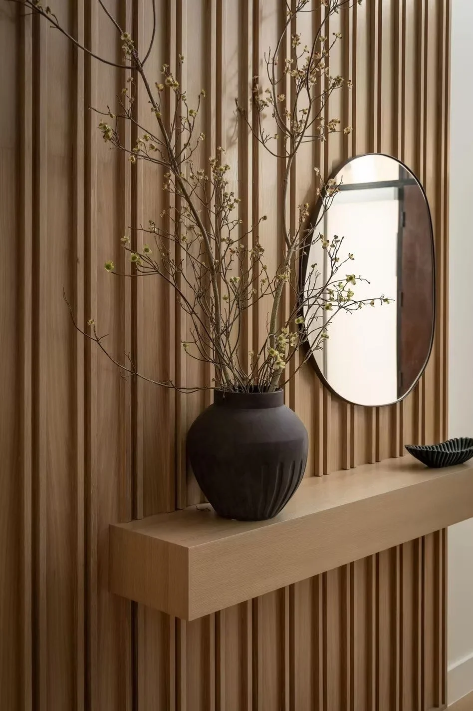 A black vase with dried branches and small yellow flowers sits on a floating wooden shelf against a slatted wood wall. A mirror and a small decorative bowl are also on the shelf.