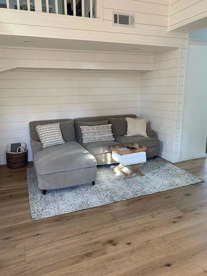 A grey sectional sofa with three decorative pillows on a textured area rug in a room with white paneled walls and wood flooring.