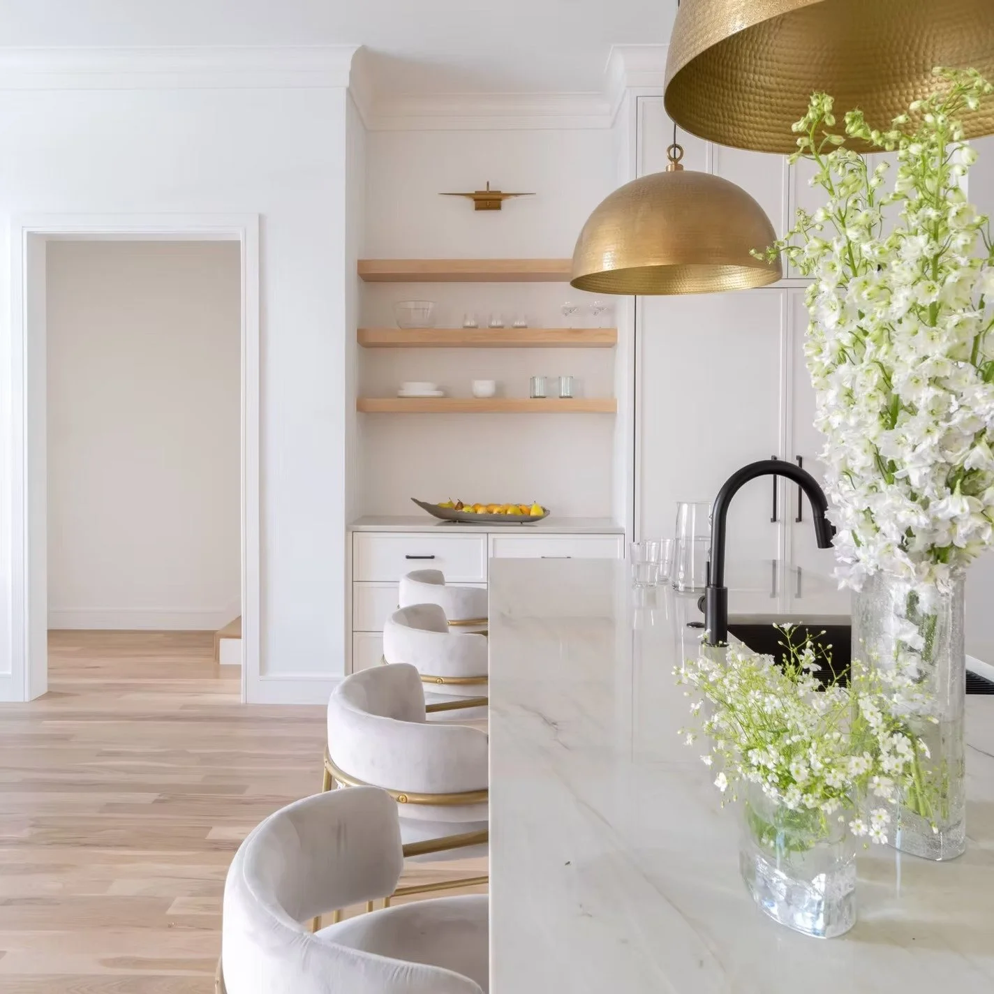 A modern kitchen with white cabinets, a marble island, and elegant chairs. There are gold pendant lights and a vase with white flowers on the marble counter, with open shelves holding glassware and bowls in the background.