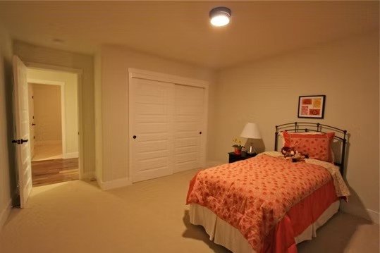 A bedroom with a bed covered in a pink patterned bedspread, a nightstand with a lamp, and a framed picture hanging above the bed.