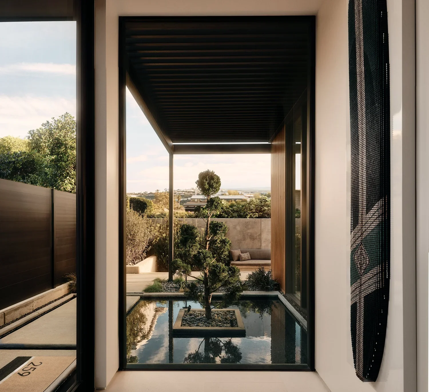 Modern balcony view with a small pond, trimmed tree, outdoor seating, and cityscape in the background, framed by glass doors.