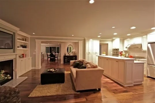 Spacious living room with a fireplace, beige sofa with pillows, black ottoman, and a large kitchen island with white cabinets, recessed lighting, and hardwood floors.