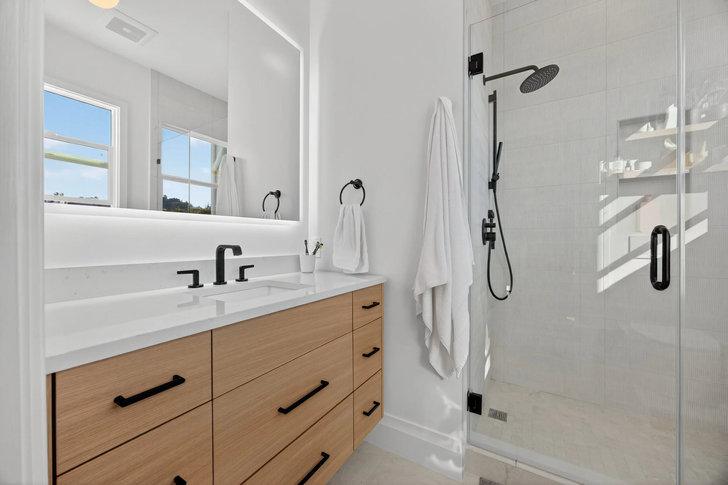 Modern bathroom with a vanity featuring a white countertop and wooden drawers, black fixtures, a large mirror, and a walk-in shower with glass door and black shower fixtures.