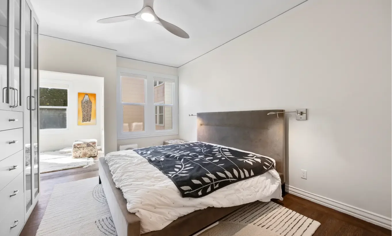 A bedroom with a bed, white walls, a window, a ceiling fan, a white wardrobe, and a small sitting area with windows and artwork.