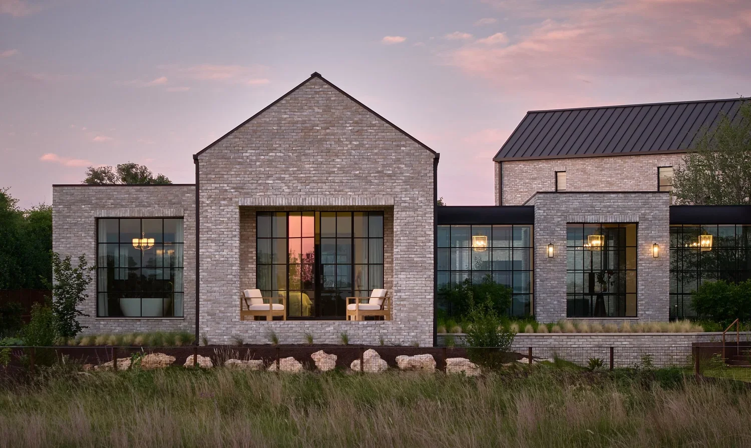 A modern house with brick exterior, large glass windows, and a dark metal roof, situated in a natural landscape during sunset.