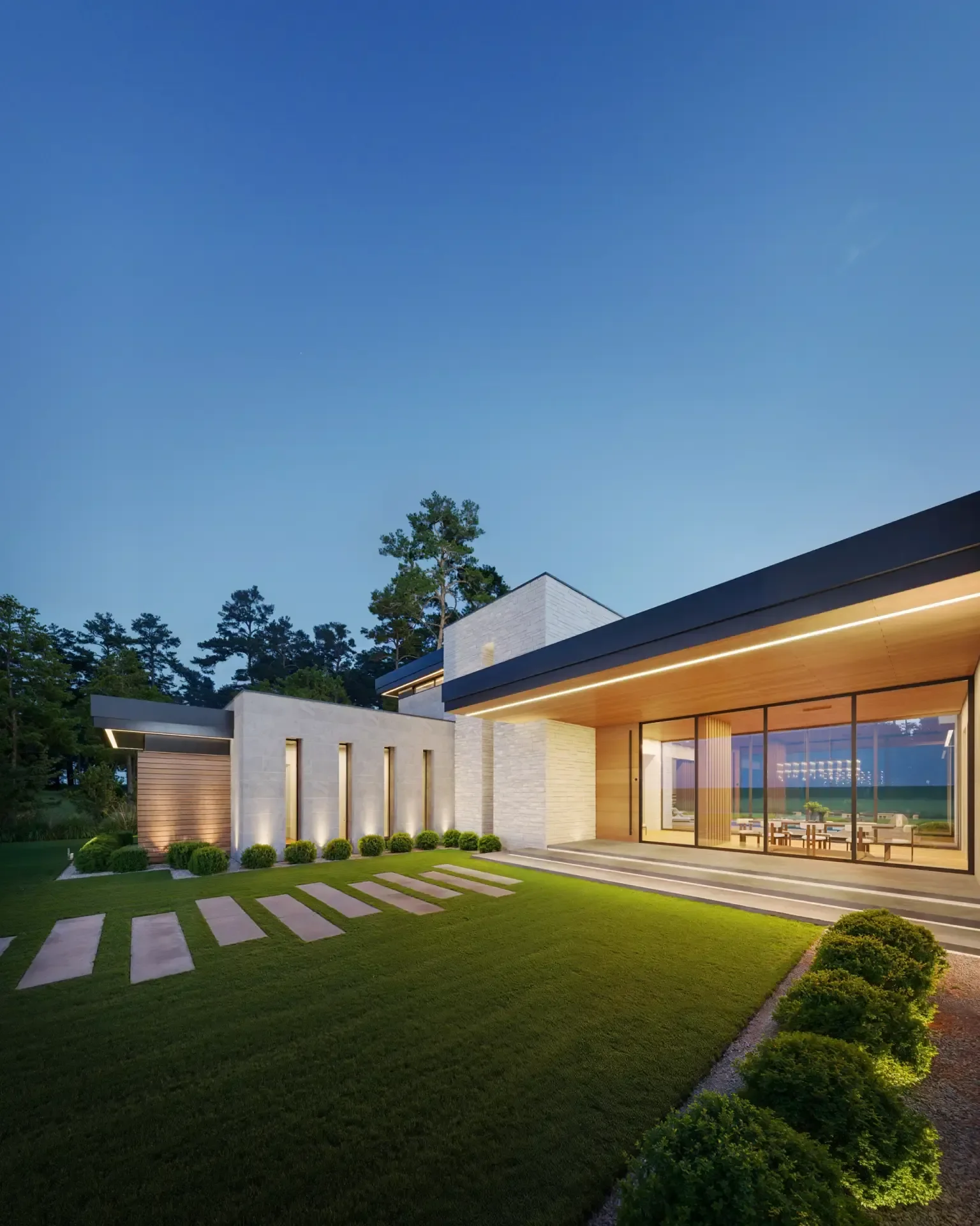 Modern house with large glass windows, wooden accents, and well-manicured lawn with stepping stones and shrubbery, illuminated by exterior lighting, under a clear evening sky.
