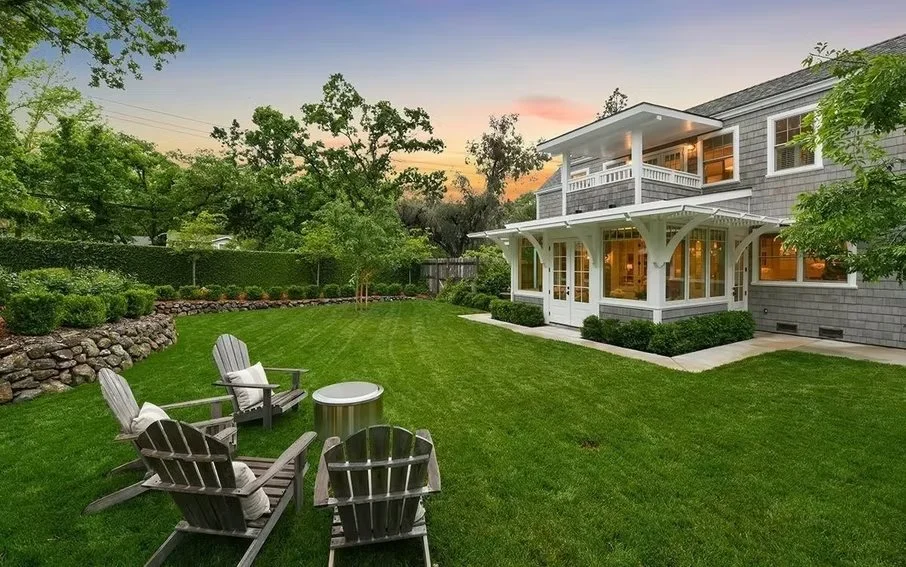 Backyard scene at sunset featuring green lawn, four wooden Adirondack chairs with pillows, a round metal table, a stone retaining wall, and a two-story house with a porch and sunroom.