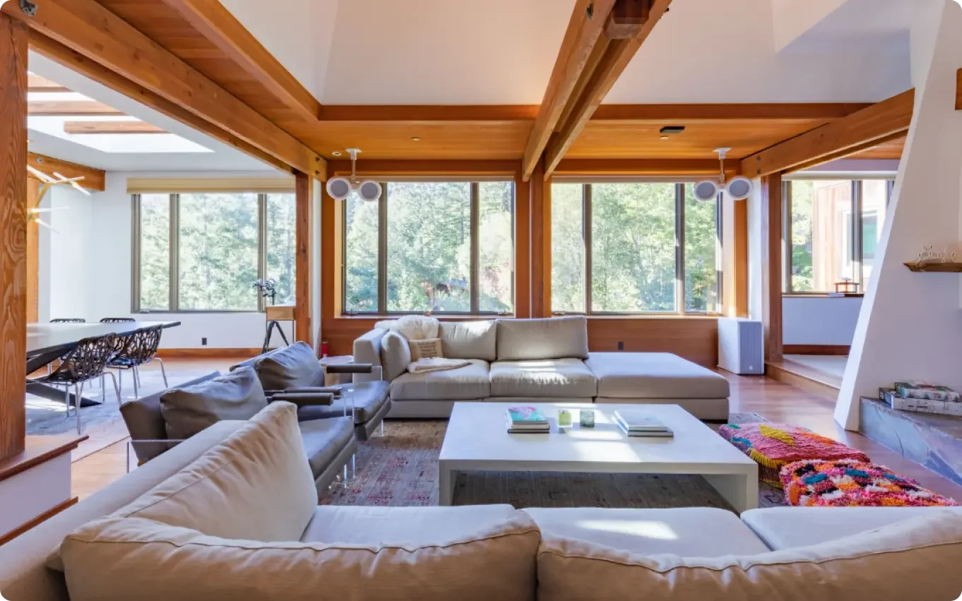 Bright living room with large windows, white and beige sofas, a white coffee table with books and candles, colorful rug, wooden beams and trim, and a dining area nearby with black chairs.