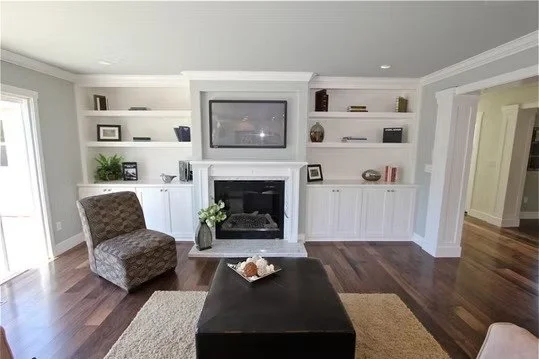 Living room with built-in white shelves around fireplace, flat-screen TV mounted above, dark wood floor, and a brown patterned armchair near the window.