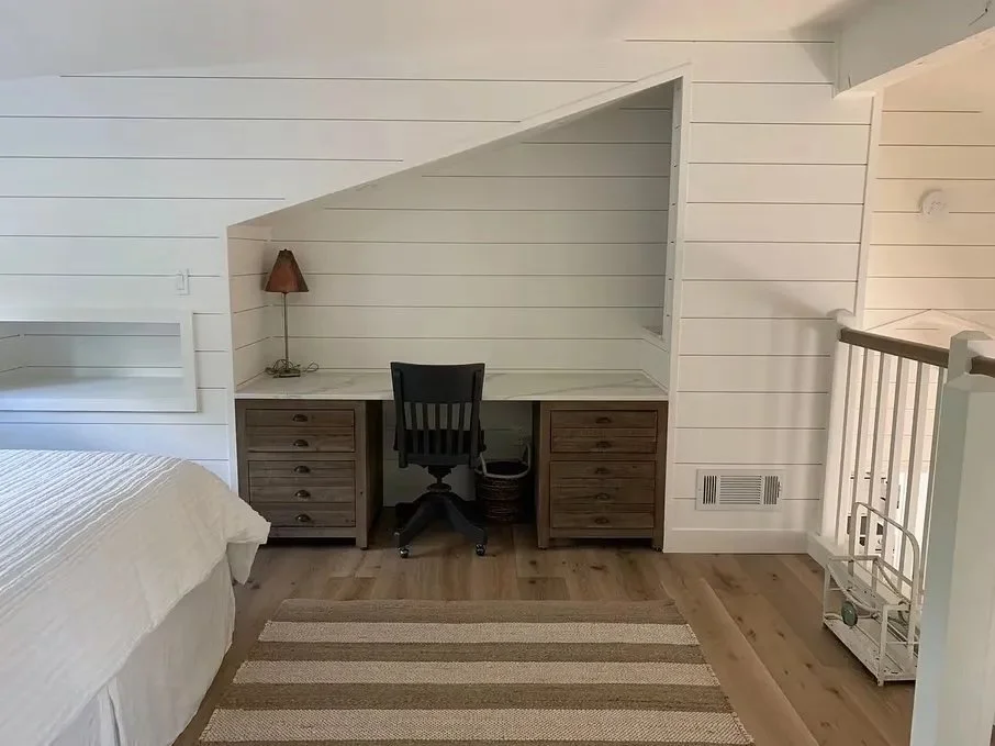 Home office desk with black chair, two wood drawers on each side, small lamp, window, and stairs with white railing in a room with white shiplap walls and wooden floor.