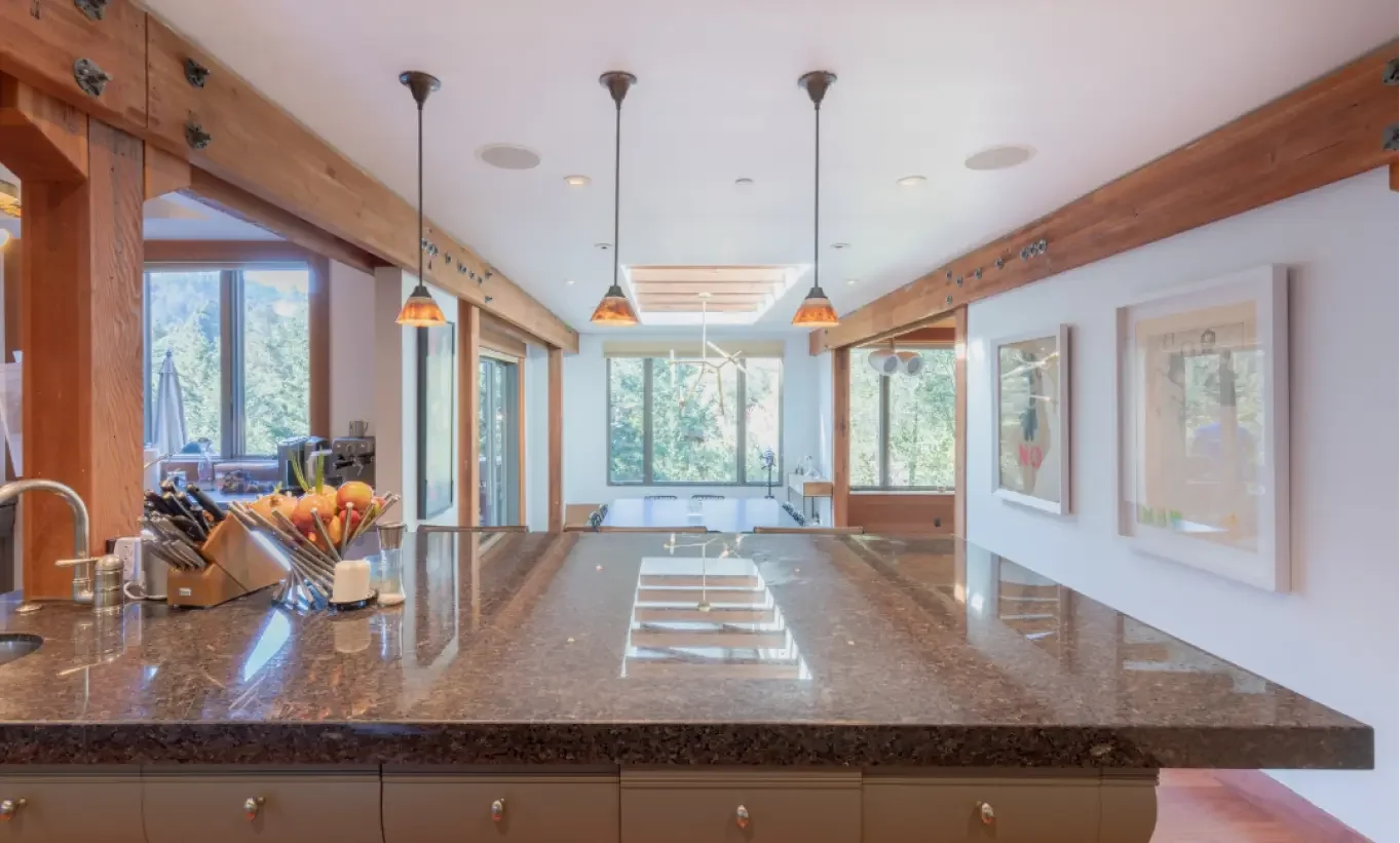 View of a kitchen island with a polished dark granite countertop, surrounded by wooden beams and windows showing a lush green outdoor scene, with framed artwork on white walls.