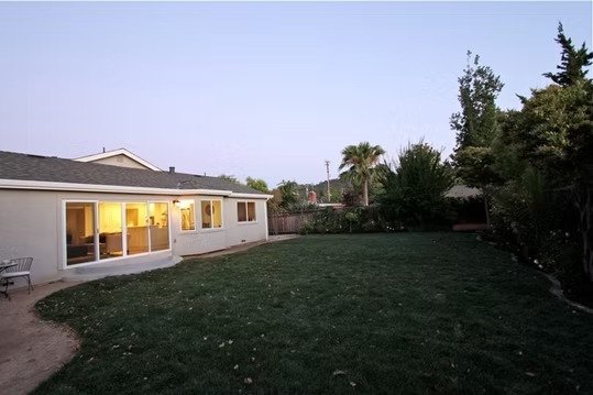A backyard with a grassy lawn, a white house with windows emitting yellow light, and various trees and bushes in the background during evening.