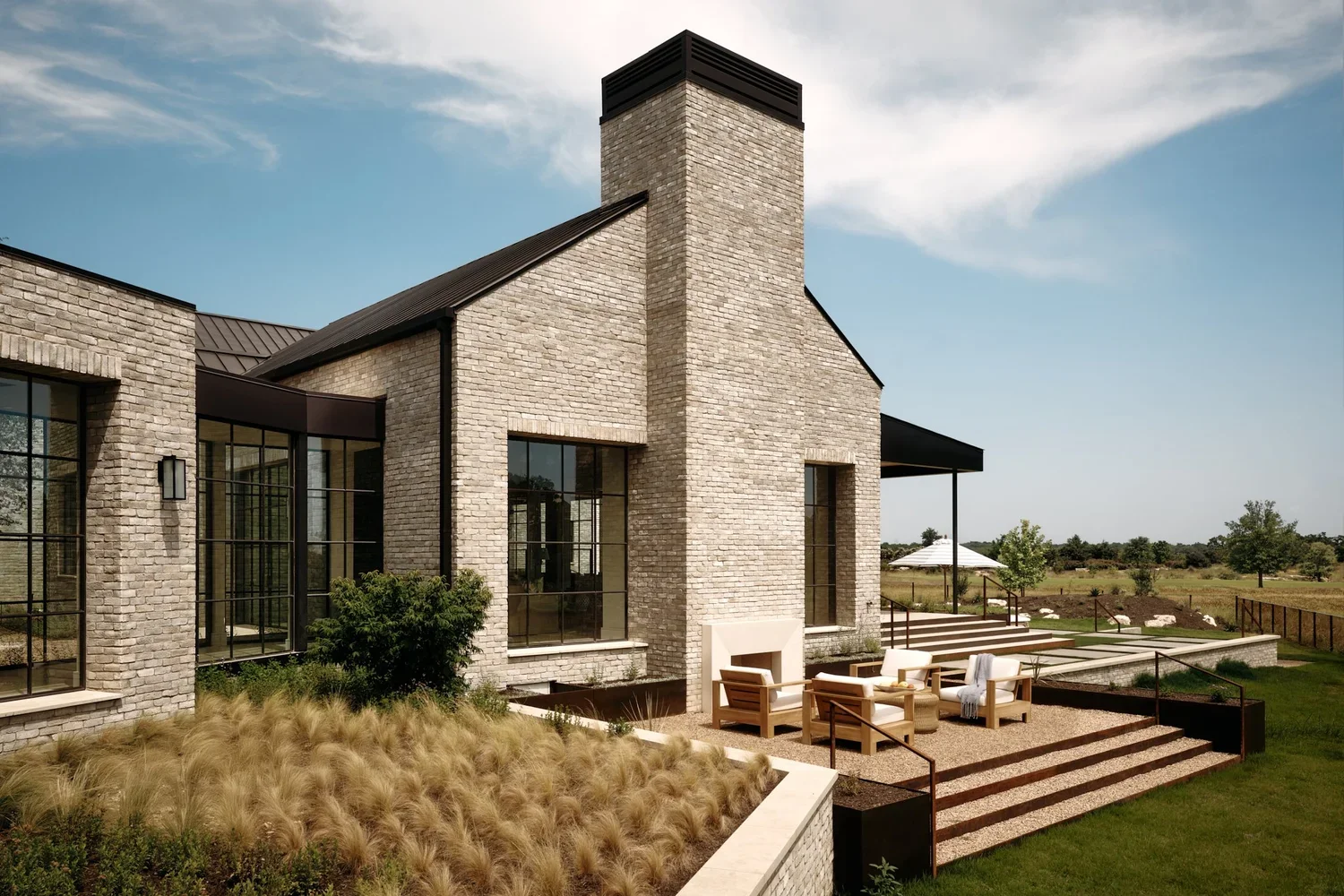 Modern brick house with large glass windows, fireplace, outdoor seating area on a patio with steps, surrounded by landscaped garden and open field under a blue sky.