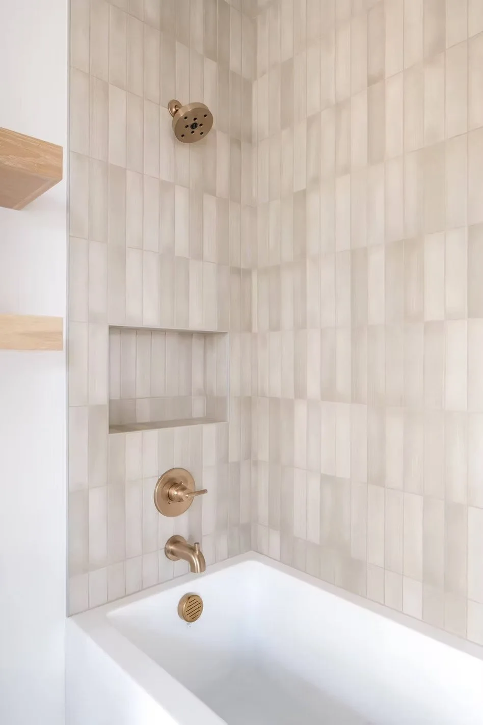 A modern shower with beige tiles, a bronze showerhead, a control handle, a built-in shelf, and a white bathtub.