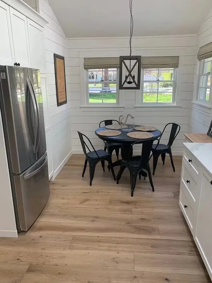 A small dining area with a round black table and four black chairs, set with round woven placemats, in a room with white wooden panel walls, three windows, and a modern chandelier hanging above the table. To the left is a stainless steel refrigerator