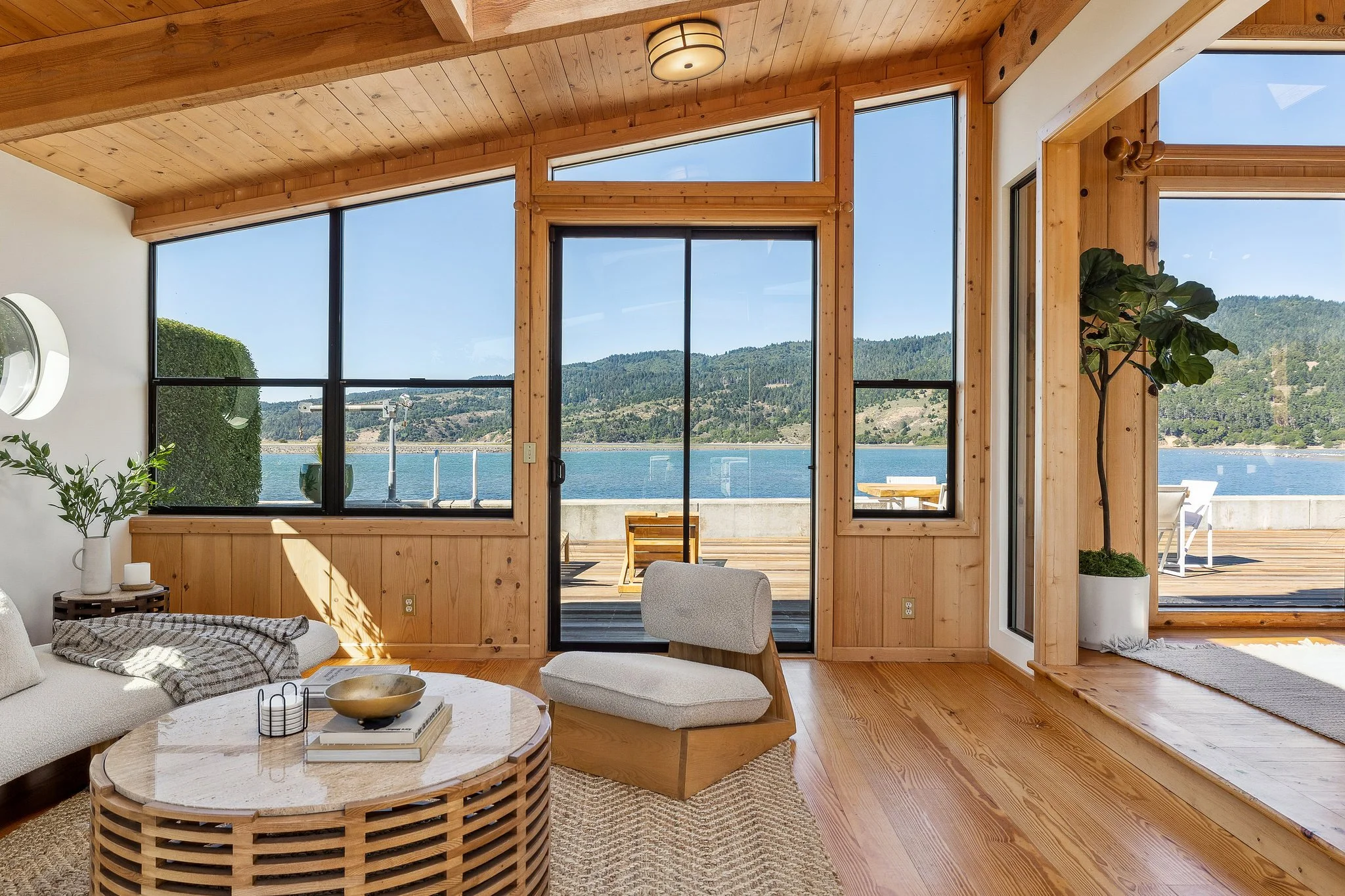 View from inside a modern home with large windows overlooking a lake, with wooden interior walls and ceilings, and a balcony outside, featuring outdoor furniture.