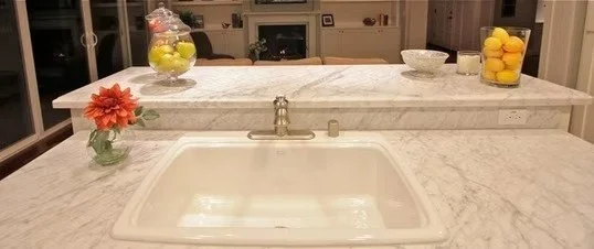 Kitchen island with a marble countertop, a white sink, and decorative fruit bowls and flowers.