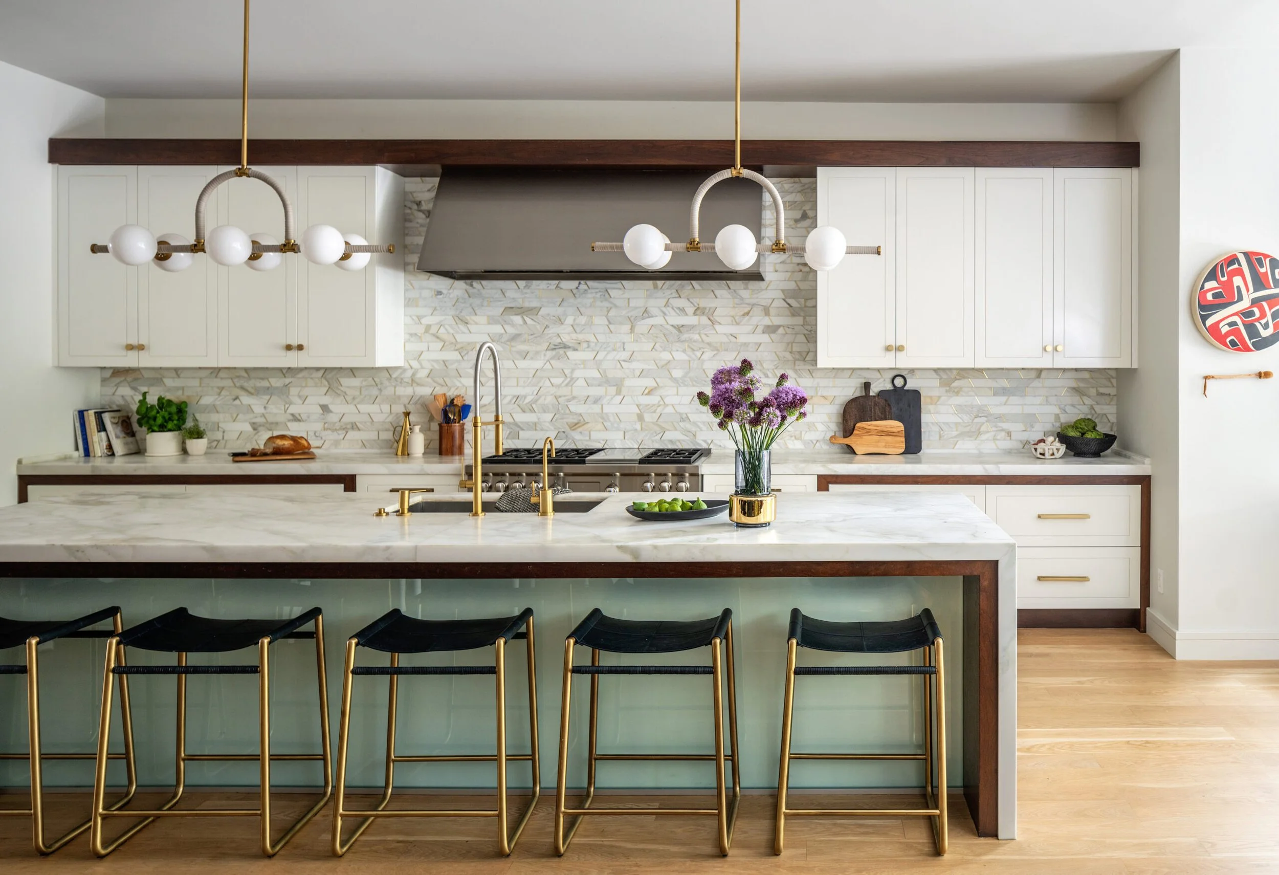 Modern kitchen with white cabinets, a marble island, gold fixtures, black chairs with gold legs, a vase of purple flowers, and a backsplash with geometric tile pattern.