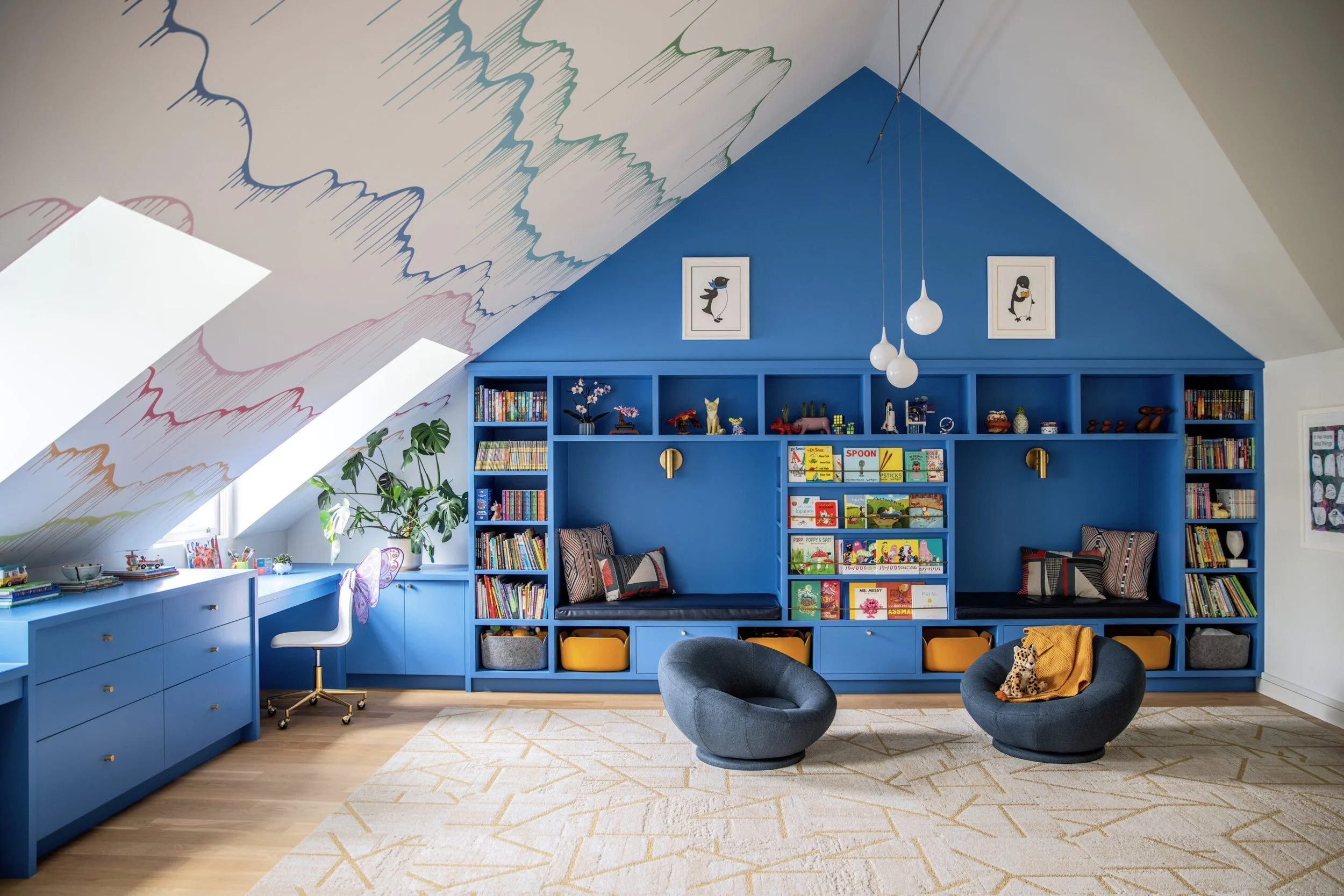 Colorful children’s library or playroom with blue built-in bookshelves, featuring toys, books, and plush animals, two gray armchairs, a white desk with a white chair, large windows, and artwork of penguins on the blue wall, with a painted cloud desig