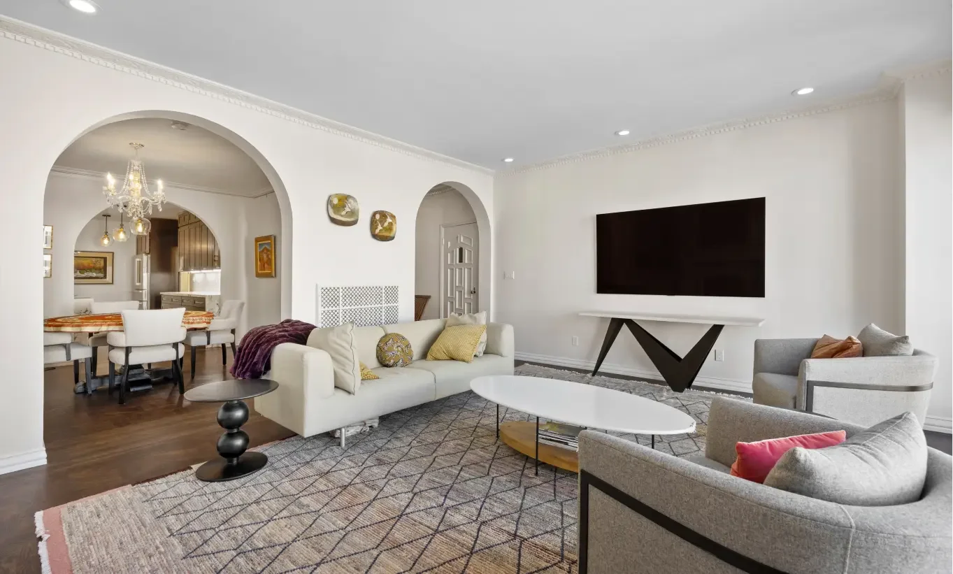 Living room with white walls, a wall-mounted large black TV, a beige sofa with pillows, and a round white coffee table. To the right, a gray armchair with a pillow. In the background, a dining area with a chandelier, wooden table, and white chairs. T