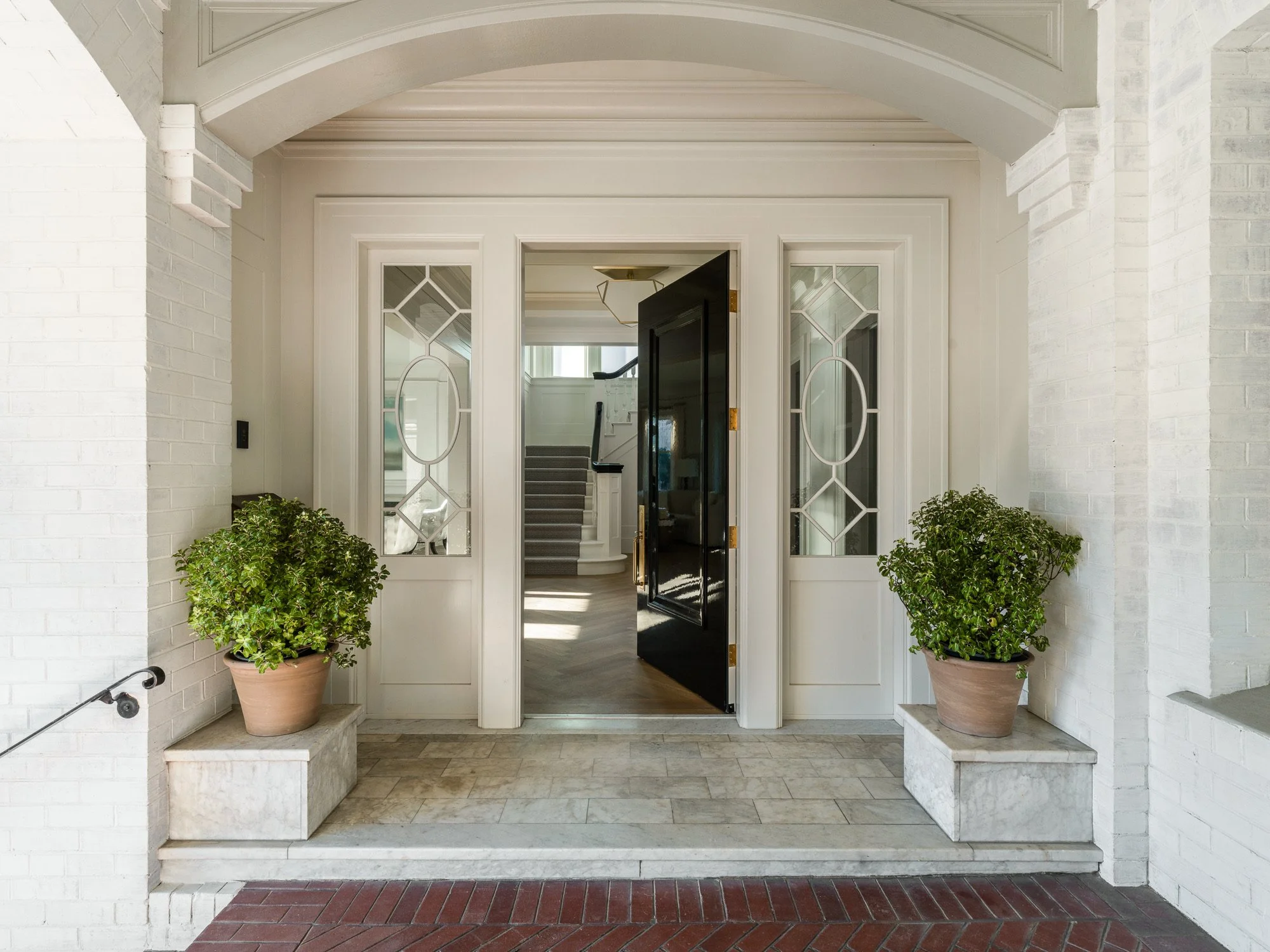 Entryway with white brick walls and large potted green plants on either side of a black front door with glass panels, leading to a staircase inside.