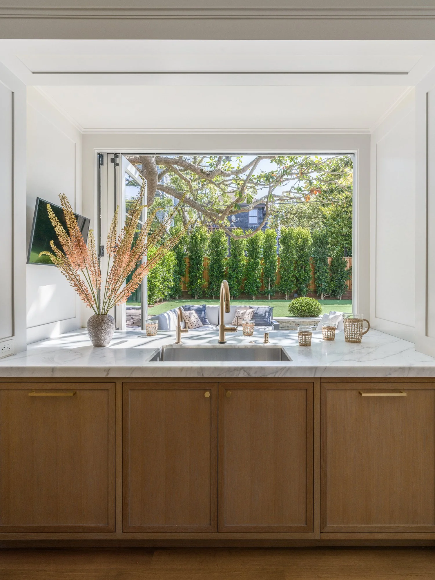 A kitchen with a marble countertop, a sink with a gold faucet, and a view of a backyard with a tree, green bushes, and outdoor furniture.