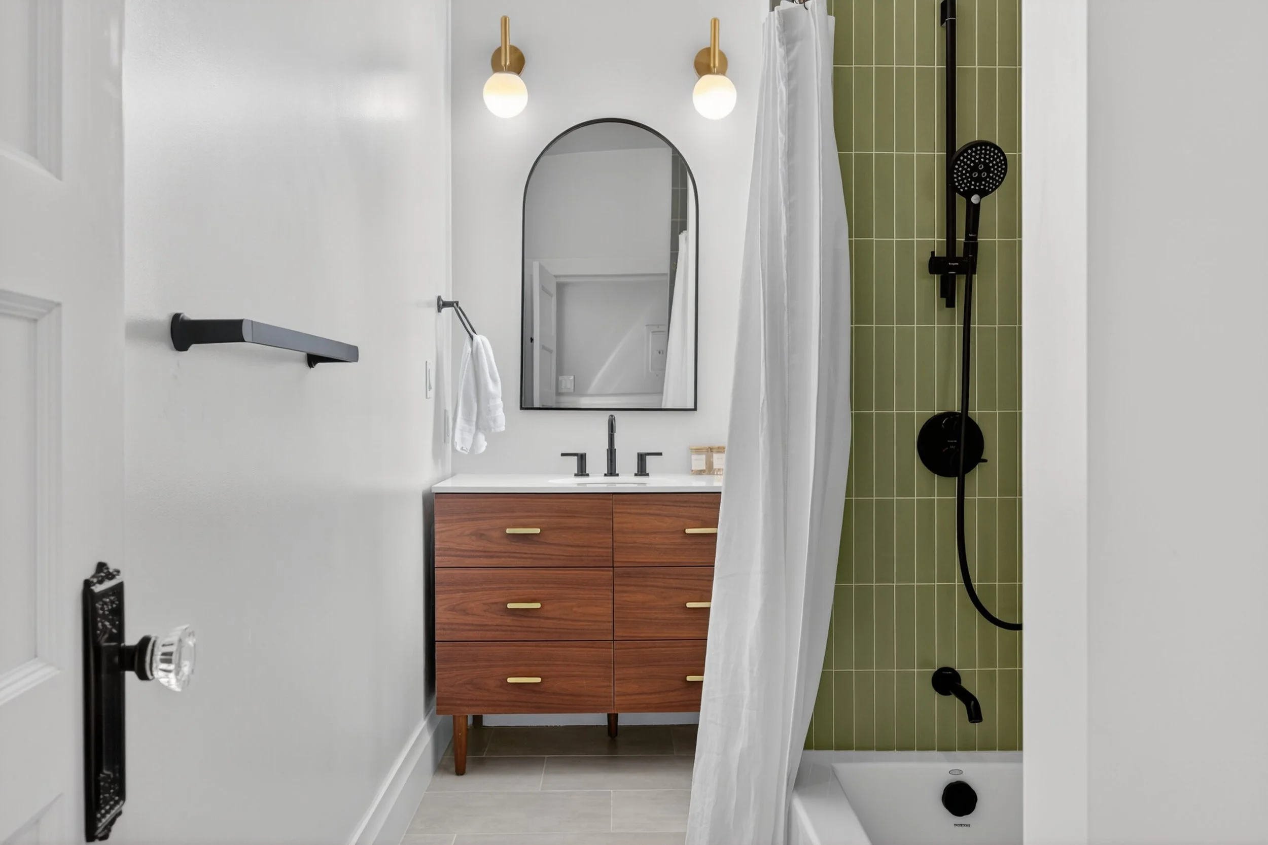 Modern bathroom with white walls, a wooden vanity with yellow handles, an oval mirror, and a shower area with a green tiled wall and black shower fixtures.