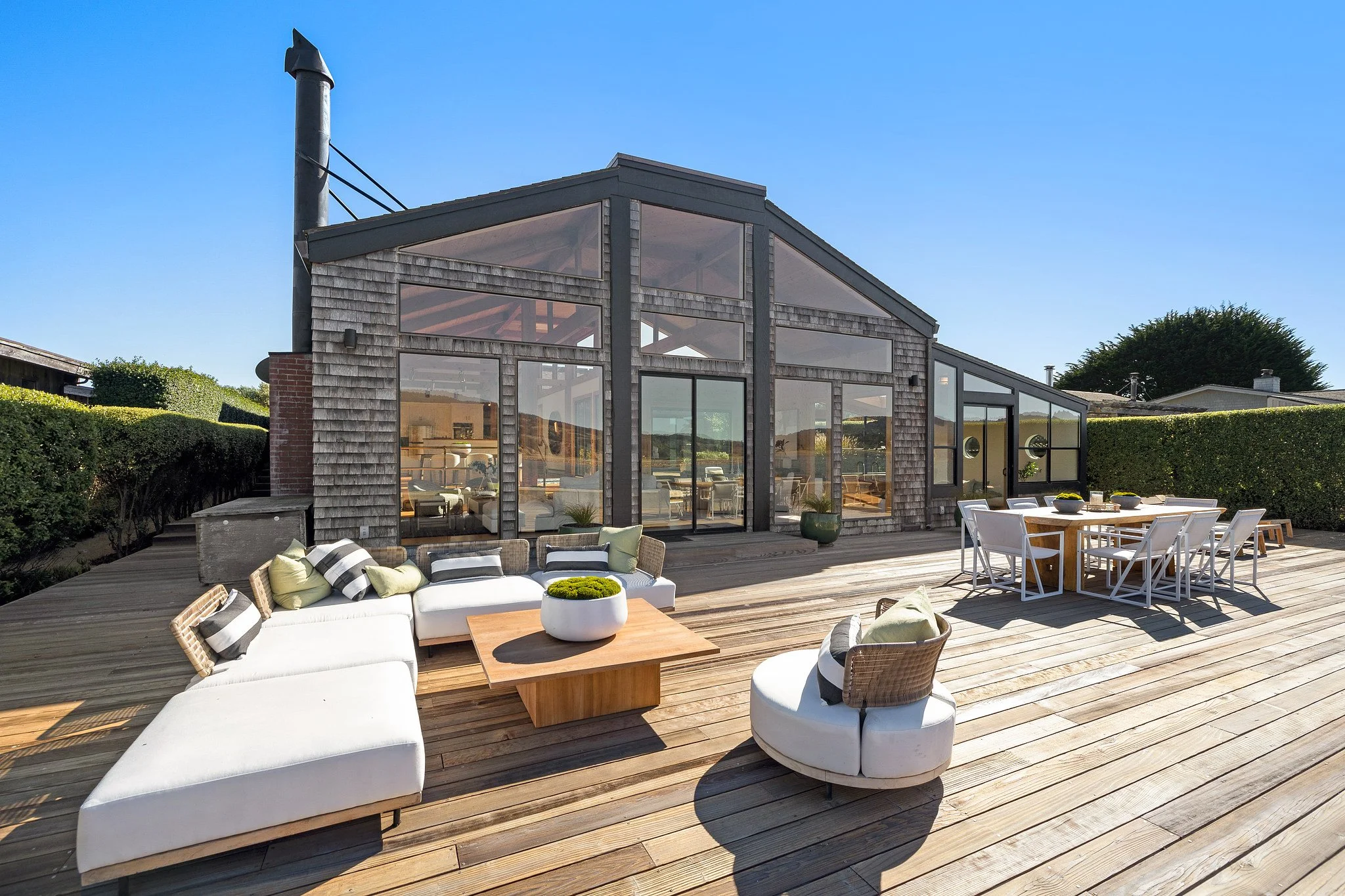 An outdoor patio with wooden decking, modern white outdoor furniture with cushions, and a large house with extensive glass windows reflecting the sky and surrounding greenery.